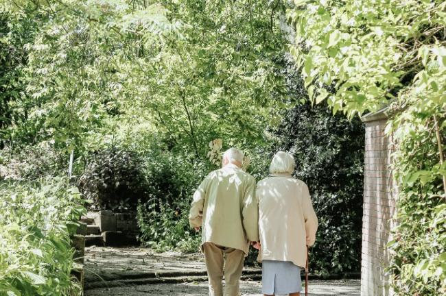 Elderly couple walking arm in arm along a shaded garden path