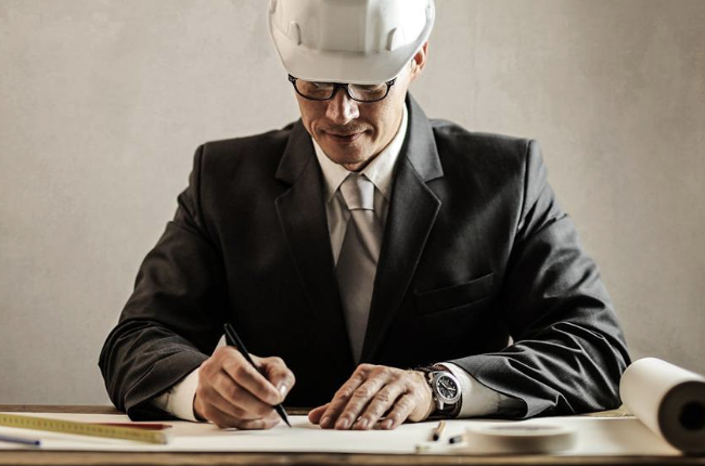 Man in suit and white hard hat drawing on blueprints at a desk