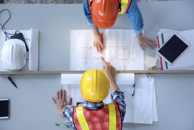 Overhead view of two construction workers in hard hats reviewing blueprints on a table