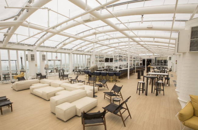 Rooftop lounge under a white-framed glass enclosure with sofas, bar and high-top tables