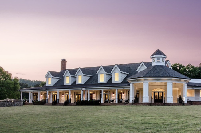 White clubhouse building with dormers, cupola and wraparound porch at dusk