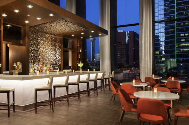Hotel bar and lounge with marble bar, stools and orange chairs beside floor-to-ceiling windows overlooking a city skyline at dusk