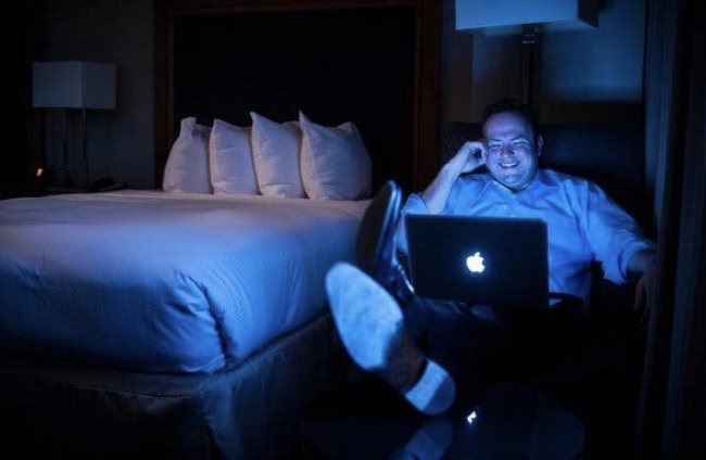 Man relaxing in a dark hotel room using a laptop beside the bed