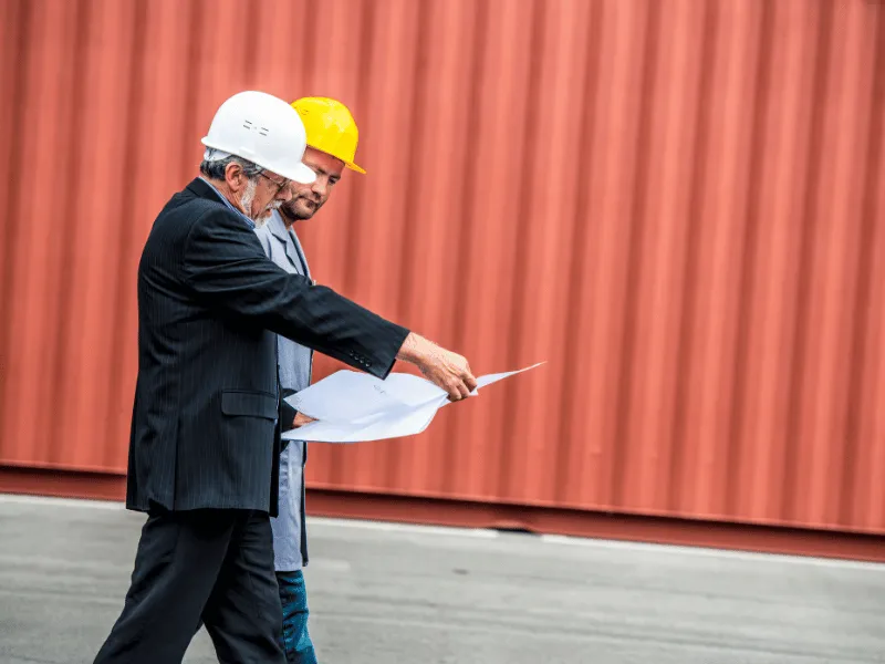 Two men in hard hats reviewing plans in front of a shipping container