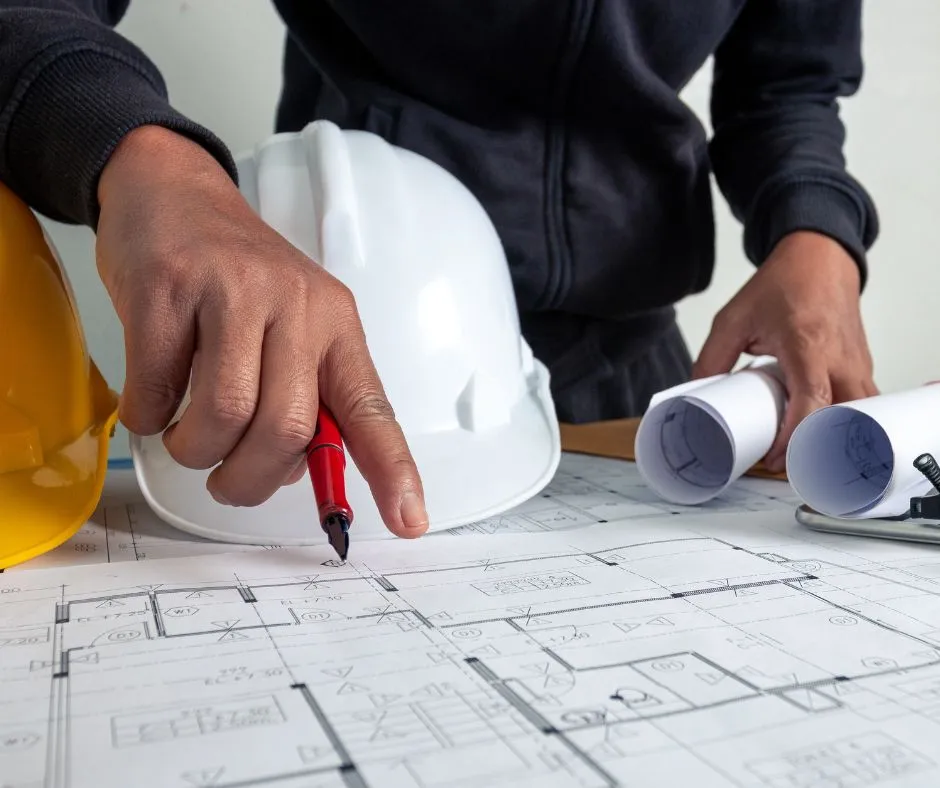 Hands pointing a pen at architectural blueprints next to hard hats