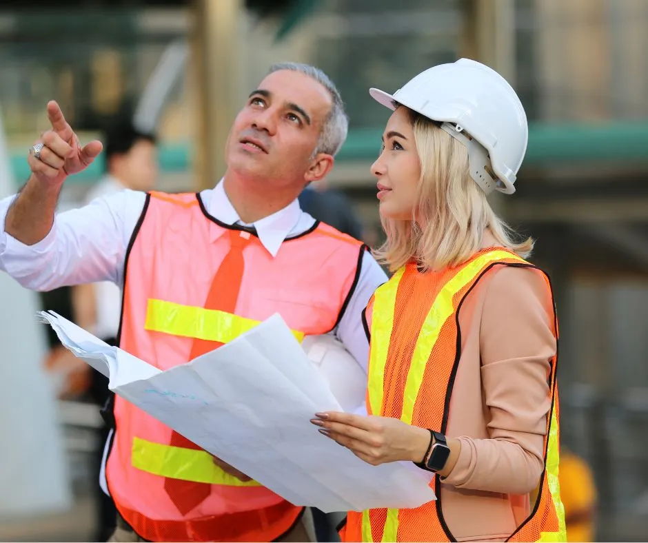 Two construction professionals in safety vests and hard hats reviewing plans on site