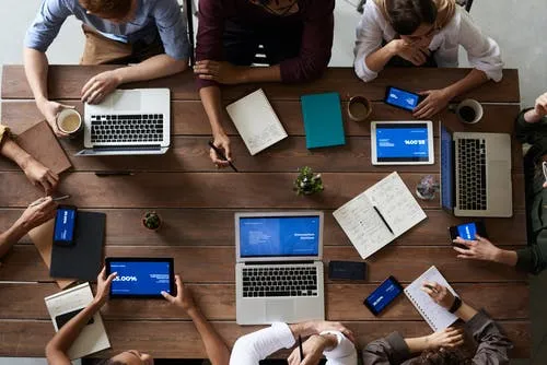 Overhead view of a team meeting around a wooden table with laptops, tablets and phones