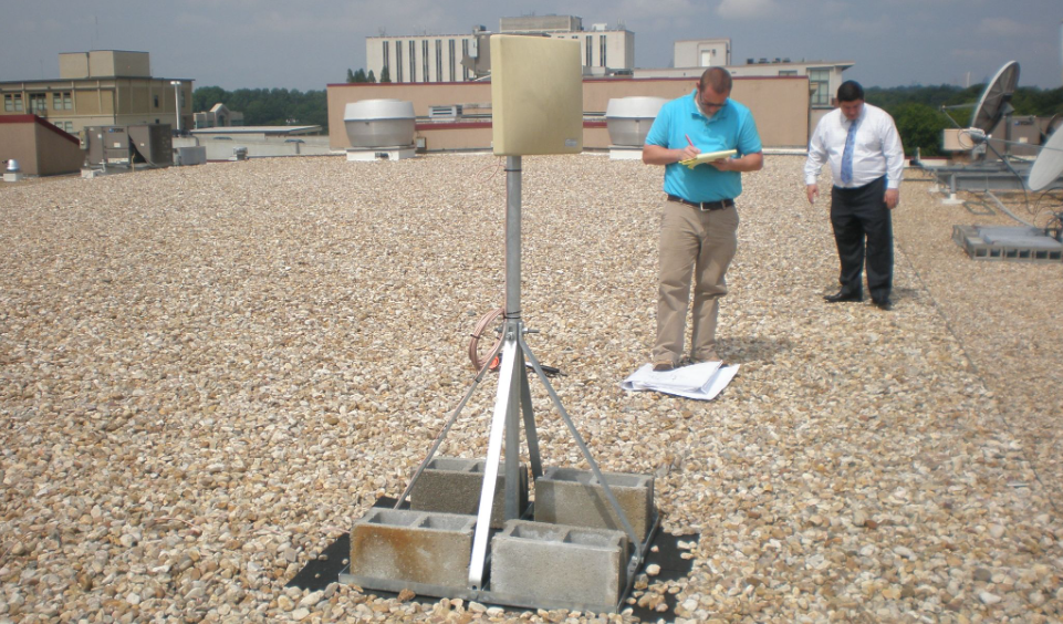 Two men surveying a gravel rooftop beside a pole-mounted antenna